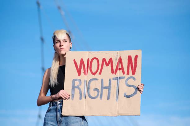 Waist-up studio portrait of a woman looking into the camera with a blank expression. She is holding a white sign displaying nothing on it.