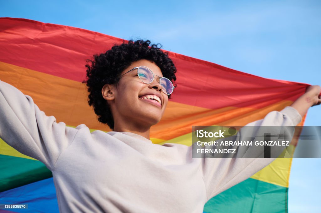 Close up view of an African American young woman holding and raising a rainbow flag over the blue sky LGBTQIA People Stock Photo