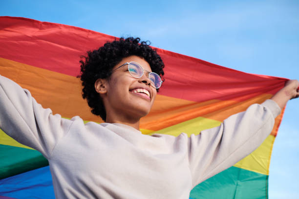 Close up view of an African American young woman holding and raising a rainbow flag over the blue sky Close up view of an African American young woman holding and raising a rainbow flag over the blue sky lgbtqia people stock pictures, royalty-free photos & images
