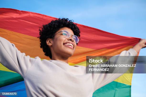 Close Up View Of An African American Young Woman Holding And Raising A Rainbow Flag Over The Blue Sky Stock Photo - Download Image Now