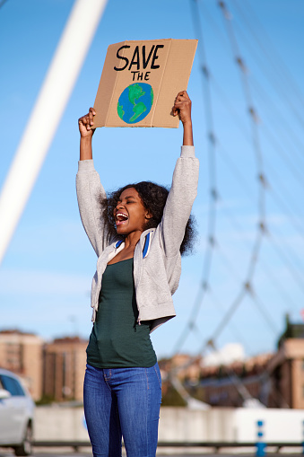 Vertical image of a young African American female protester with curly hair shouting as she raises her arms holding a banner and protesting against environmental pollution.