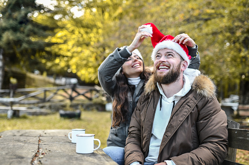 Smiling Female Setting Up Boyfriend's Christmas Hat While Enjoying Tea In Forest