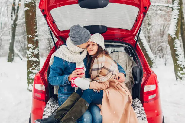 Photo of Couple in love sitting in car trunk drinking hot tea in snowy winter forest. People relaxing outdoors during road trip