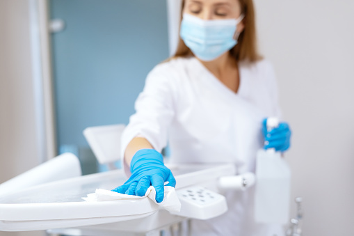 Nurse in protective gloves cleaning work surface at stomatology clinic, sanitizing table with disinfectant spray bottle, washing dental chair with towel, selective focus