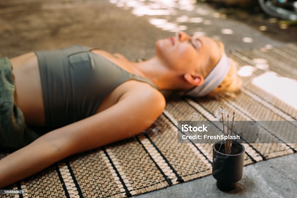 Woman Practicing Yoga with Incense Sticks beside her An anonymous female lying down on a rug and meditating outdoors, focus on incense sticks. Yoga Stock Photo