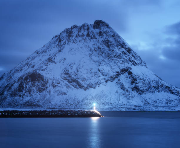 Lighthouse, sea and beautiful snow covered mountain and cloudy blue sky reflected in water at dusk. Winter landscape with lighthouse, snowy rock at night. Lofoten islands, Norway at twilight. Nature Lighthouse, sea and beautiful snow covered mountain and cloudy blue sky reflected in water at dusk. Winter landscape with lighthouse, snowy rock at night. Lofoten islands, Norway at twilight. Nature lighthouse lighting equipment reflection rock stock pictures, royalty-free photos & images