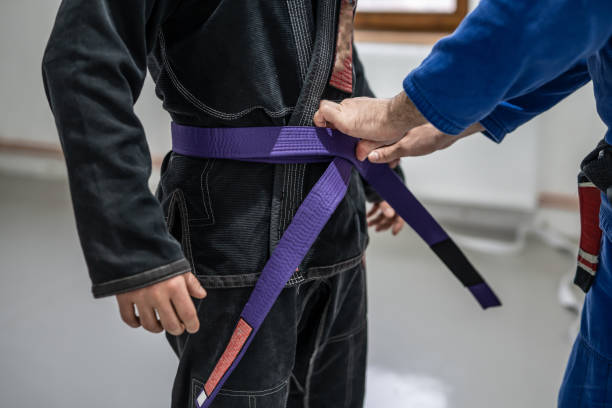 bjj brasiliano jui jutsu promozione cintura primo piano sulle mani dell'istruttore sconosciuto cintura nera professore legare cintura viola sulla vita del suo studente - purple belt foto e immagini stock