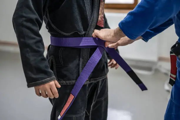 BJJ brazilian jui jutsu belt promotion close up on hands of unknown instructor black belt professor tie up purple belt on waist of his student