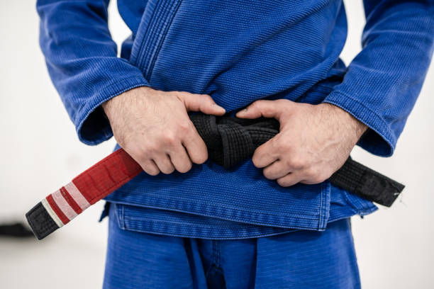 Close up on midsection of unknown caucasian male athlete bjj brazilian jiu-jitsu black belt standing on the mat at academy holding his belt during training wearing blue gi front view Close up on midsection of unknown caucasian male athlete bjj brazilian jiu-jitsu black belt standing on the mat at academy holding his belt during training wearing blue gi front view brazilian jiu jitsu photos stock pictures, royalty-free photos & images