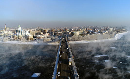 The old bridge over the Angara river. Steam is emitted from the water. Aerial drone flight. In winter, the Russian city is covered with ice and snow.