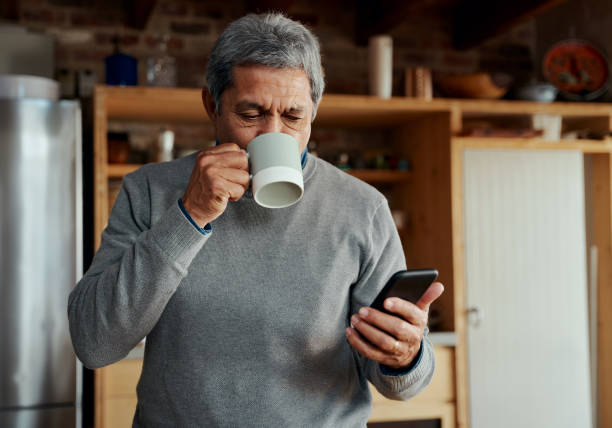 homem idoso multicultural tomando um gole de café da manhã enquanto lê uma mensagem no smartphone na cozinha moderna - senior adult human face male action - fotografias e filmes do acervo