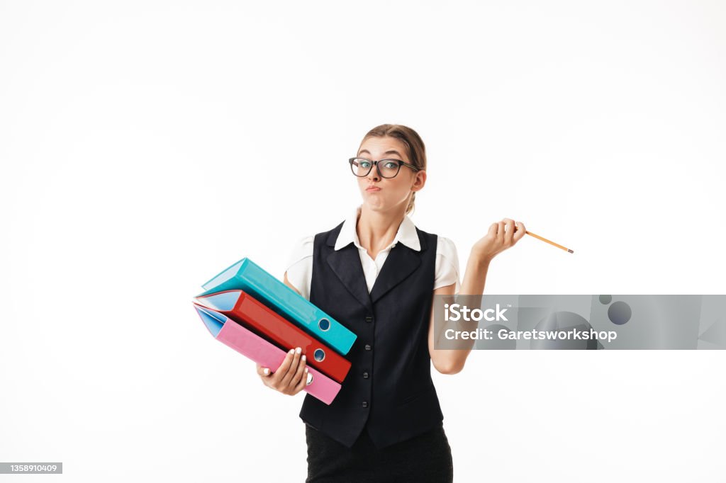 Young woman in black vest and eyeglasses thoughtfully looking in camera while holding colorful folders and pencil in hands over white background Young woman in black vest and eyeglasses thoughtfully looking in camera holding colorful folders and pencil in hands over white background Busy Stock Photo