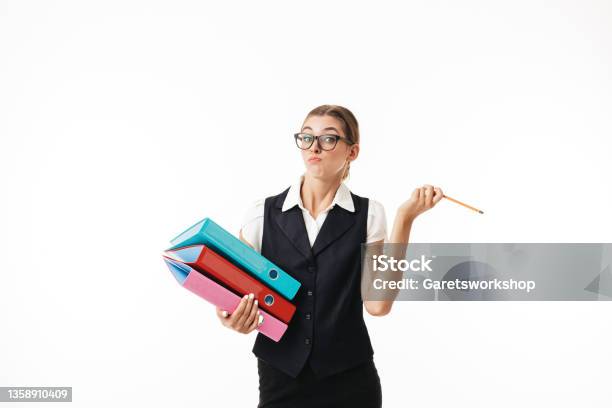 Young Woman In Black Vest And Eyeglasses Thoughtfully Looking In Camera While Holding Colorful Folders And Pencil In Hands Over White Background Stock Photo - Download Image Now