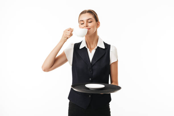 istock Young beautiful waitress in uniform holding tray while smelling fresh cup of coffee  over white background 1358910414