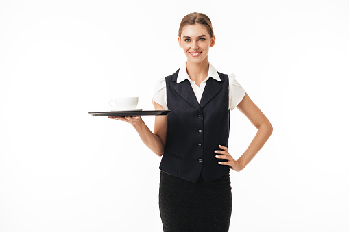 Young smiling waitress in uniform holding tray with cup of coffee dreamily looking in camera over white background