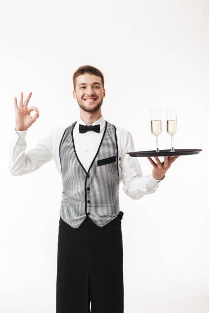 istock Young smiling waiter in uniform joyfully looking in camera holding tray with glasses while showing ok gesture over white background 1358910429