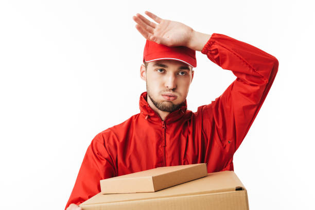 Young delivery man in red cap and jacket holding boxes in hands tiredly looking aside over white background