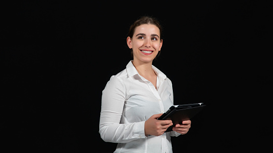 Young business woman looking at her black tablet. She studies the developments in technology with her digital tablet. Studio shot of career man in white shirt. Woman looking up and smiling. Successful person is quite happy.