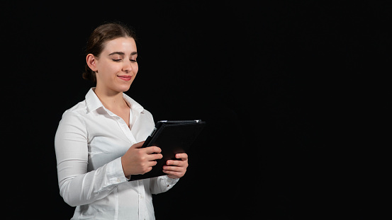 Standing teenage girl in a white shirt with her hair down is broadcasting with her tablet. The phenomenon speaks to its female followers. He gives education and carries out activities in the social field. Social gathering is happening. The beautiful lady is talking to her followers on the tablet. There is a woman in front of a black background.