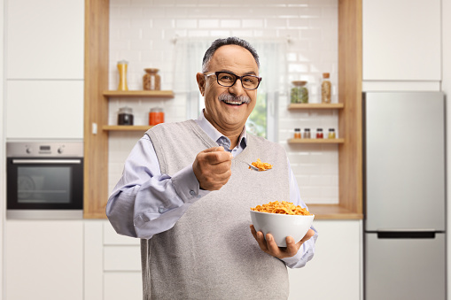Smiling mature man standing in a kitchen and eating cornflakes