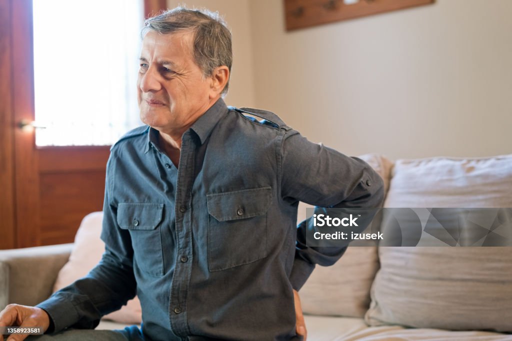 Elderly man suffering from backache at home Senior man having backache sitting at home. Elderly male is suffering from pain. He is wearing casuals. Hip - Body Part Stock Photo