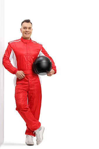 Full length portrait of a racer in a red suit leaning on a wall and holding a black helmet isolated on white background