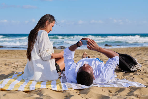 istock Young multicultural couple lying on the beach using smartphones 1358924389