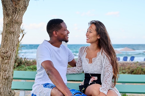 Young multicultural couple smiling talking sitting on bench, sea background. Beautiful asian woman and african american man together outdoor, sea vacation together, love, relationship, communication