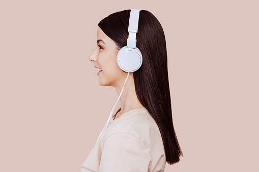 Beautiful young brunette woman girl in beige shirt posing isolated on beige wall background studio portait. Listening music with headphones. People lifestyle concept. Mock up copy space, side view.