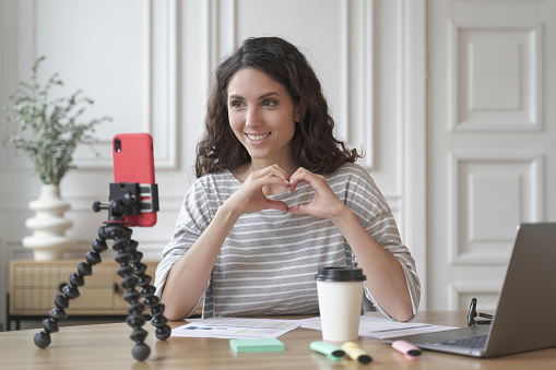 Blogging concept. Young pleasant Italian woman blogger influencer sitting in front of smartphone on tripod and shooting video, female vlogger showing heart sign, communicating with followers online