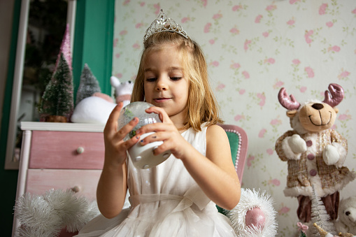 Cute little girl wearing tiara. Sitting on beautiful chair and playing with christmas ornament