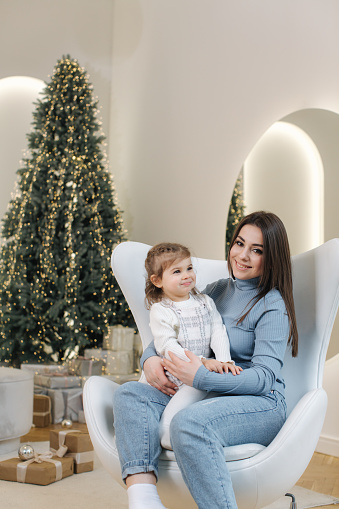 Young mom with her adorable little daughter at Chrisstmas phosession. Happy girls in front of fir tree at home. Christmas fairy tail.