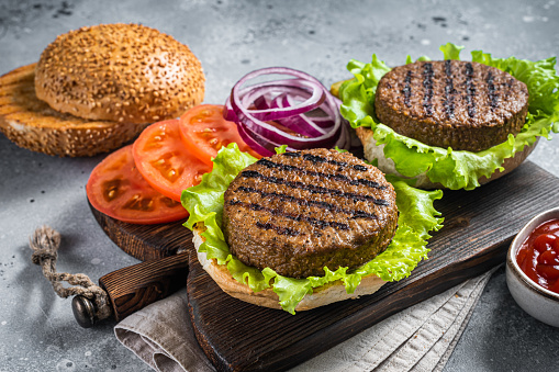 Plant based meatless burgers with vegan grilled pattie, tomato and onion on a wooden serving board. Gray background. Top view