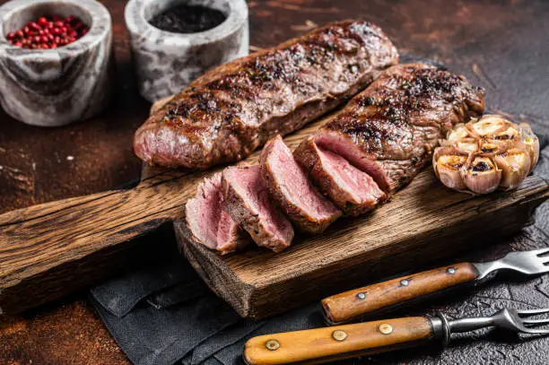 Photo of Grilled Lamb sirloin fillet meat on wooden board with herbs, mutton loin steak. Dark background. Top view. Copy space