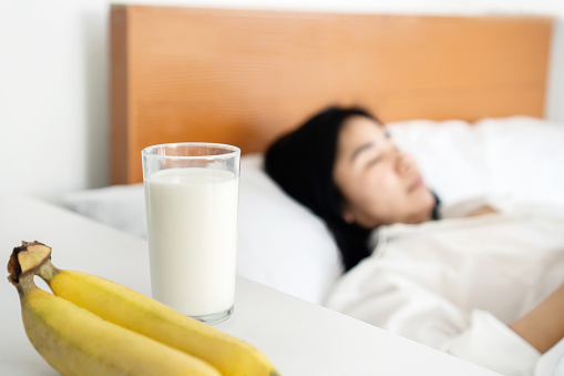 a glass of milk and bananas on the table with blur background of woman sleeping in bed