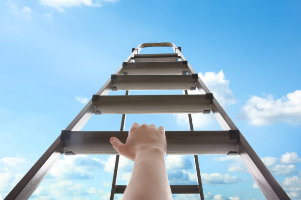 istock Woman climbing up stepladder against blue sky with clouds, closeup 1358943334