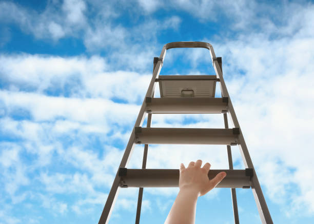 istock Woman climbing up stepladder against blue sky with clouds, closeup 1358944075