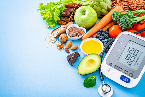 Overhead view of a blue background filled with multicolored healthy food. A blood pressure monitor and stethoscope complete the composition. The composition is at the right of an horizontal frame leaving useful copy space for text and/or logo at the left. Food included in the composition is: carrots, green apple, tomatoes, artichoke, celery, blueberries, olive oil, dates, oat, avocado, broccoli, pinto beans, chocolate, flax seeds, spinach, nuts and chia seeds among others. High resolution 42Mp studio digital capture taken with SONY A7rII and Zeiss Batis 40mm F2.0 CF lens