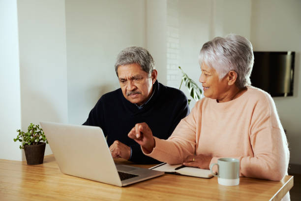istock Multi-cultural elderly couple sitting at modern kitchen counter with laptop. Wife explaining to husband 1358945564