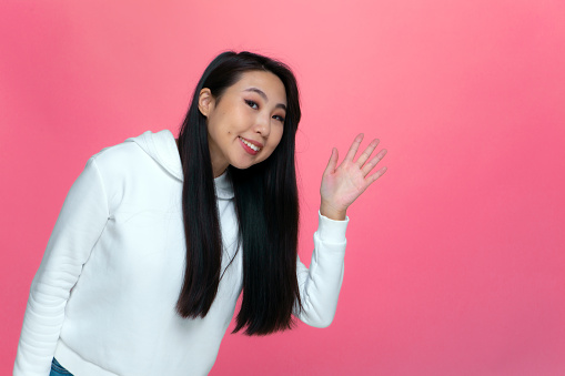 Welcome gesture. Smiling happy asian young girl blogger wave hand say hello to audience, subscribers on pink studio background. High quality photo