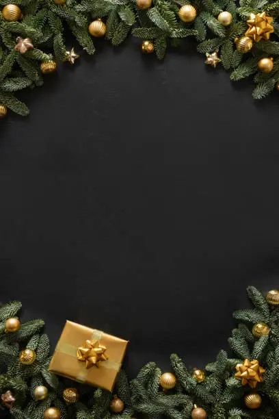 Photo of Christmas vertical frame with golden gift, baubles, evergreen branches on black background. Xmas greeting card.