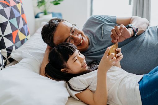 Image of an Asian Chinese young girl holding smartphone and watching video or playing games with her father on the bed