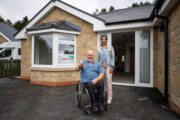 Keys to Our New Place A mid-adult couple outside of the bungalow they have just bought in Northumberland, a sold sign is in the front of the window. The man is holding up a new set of keys for their home while they are looking at the camera and smiling. The man is a wheelchair user. accessibility for persons with disabilities photos stock pictures, royalty-free photos & images