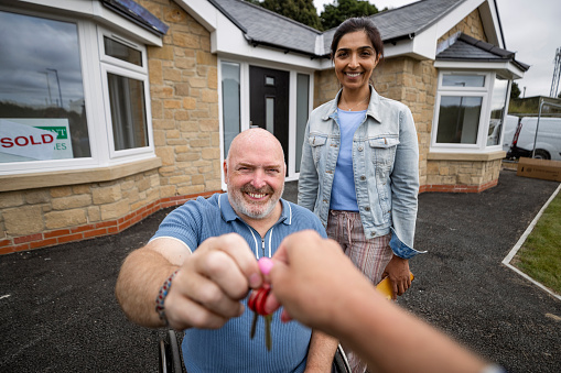 A personal perspective shot of a real estate agent passing the new house keys to a mid-adult couple outside of the bungalow they have just bought in Northumberland, a sold sign is in the front of the window. The man is a wheelchair user.