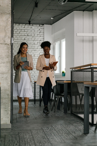 Full length shot of two young businesswomen going on a meeting, walking through a modern office space and chatting casually.
