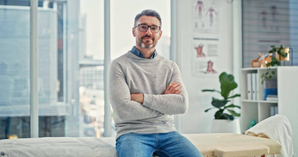Shot of a mature chiropractor sitting alone in his clinic with his arms folded stock photo