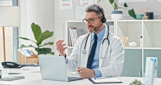 Shot of a mature doctor sitting alone and using his laptop while wearing a headset during an online consultation stock photo