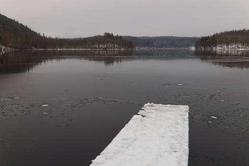 Image presents a wooden bridge or platform going towards empty lake. Winter time. Some hills in the background. Perfect reflection in the water. Plenty of copy space. Calm and quiet. Landscape of a lake in Halmstad municipality.