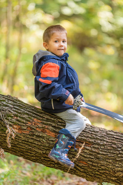 istock Young boy is sitting on Fallen Down Tree Trunk and play with a sword 1358958027