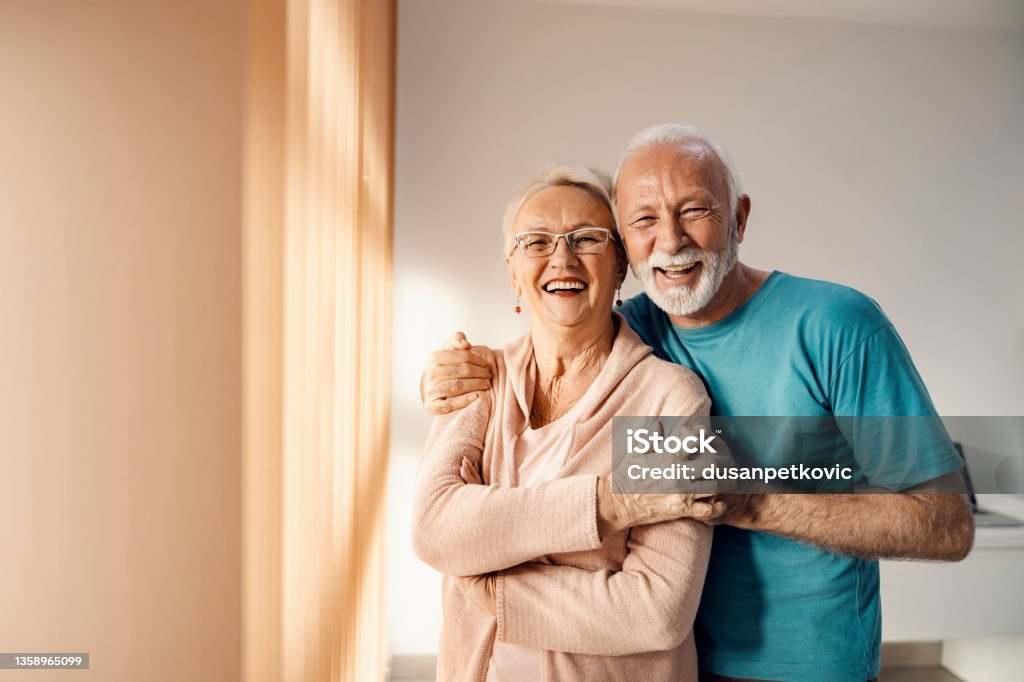Senior couple hugging in a nursing home. A happy senior couple standing next to a window in a nursing home, hugging and smiling. They have all care they need. Senior Adult Stock Photo